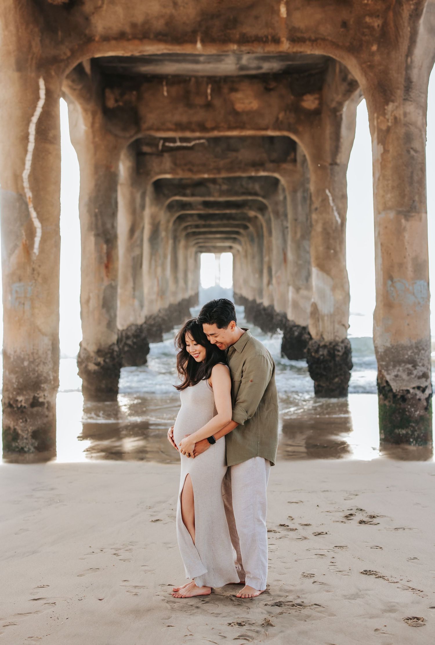 A couple embraces under a pier on the beach with geometric archways creating a stunning symmetrical backdrop.