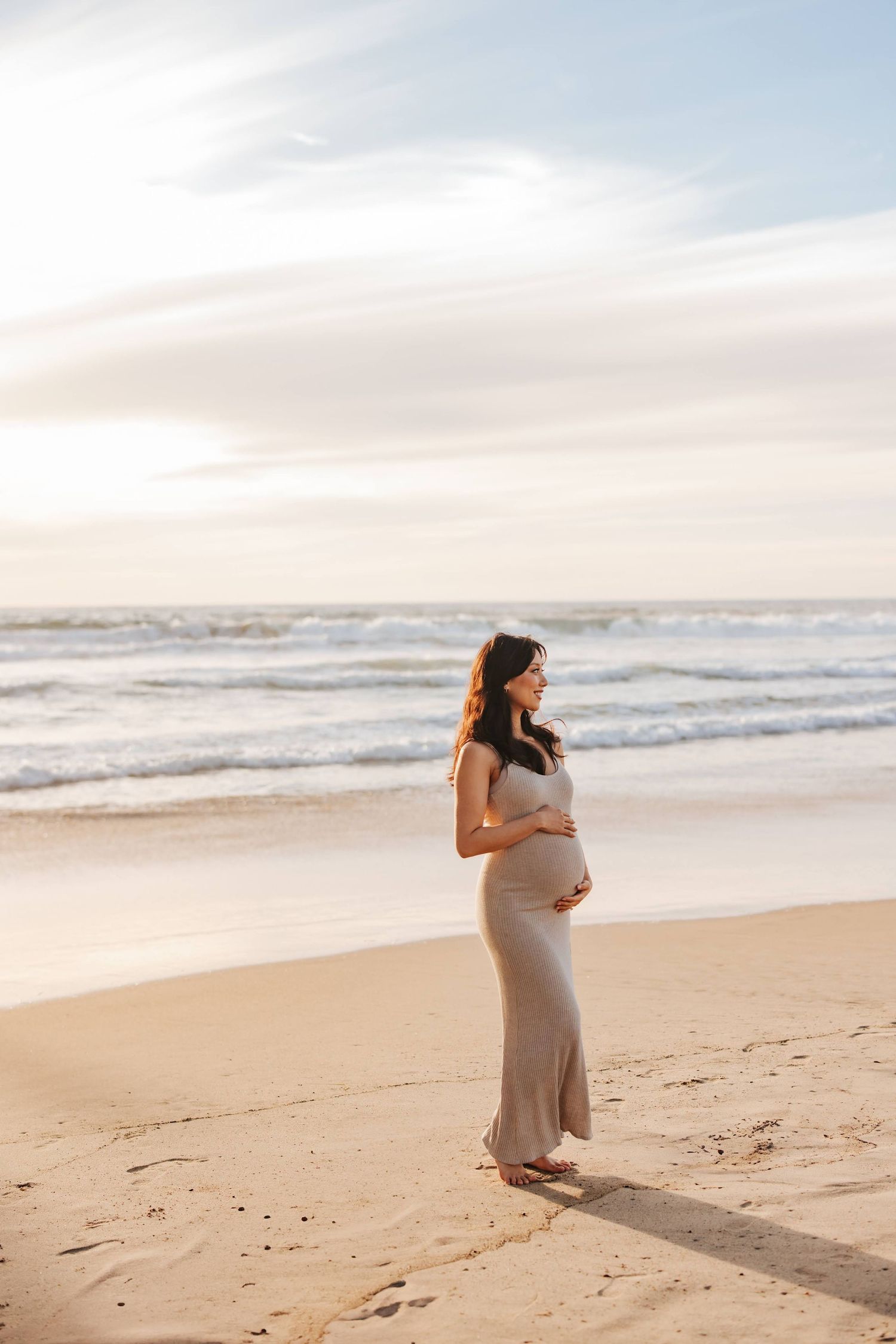 A pregnant woman in a beige maxi dress stands on a sandy beach at sunset looking out at the ocean waves.