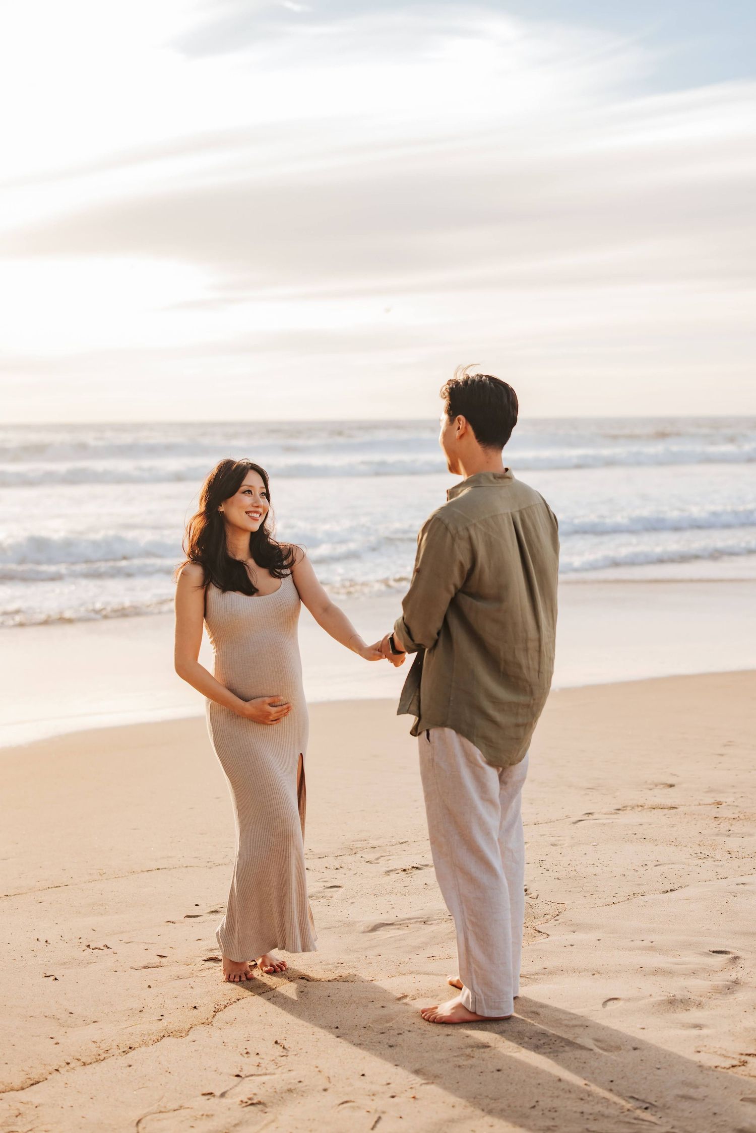 A couple holds hands while standing on a sandy beach at sunset with gentle waves in the background.