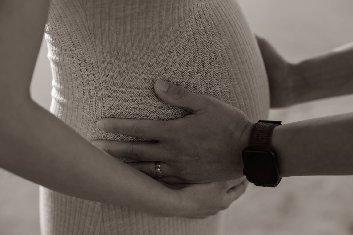 A black and white closeup of hands cradling a pregnant belly in a ribbed sweater.