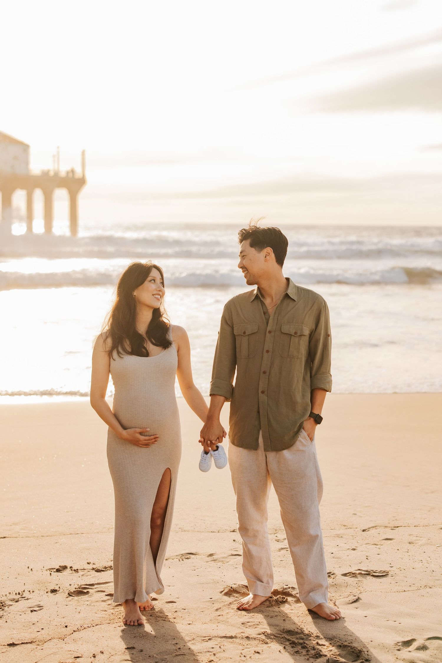 Expecting couple holding hands walking on beach at sunset with pier in background and baby shoes in hand.
