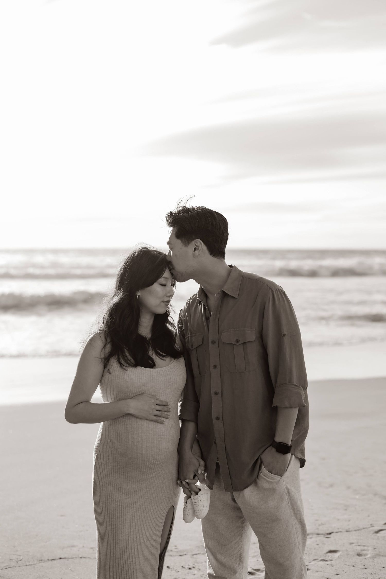 A couple shares an intimate moment on a beach at sunset, with the woman wearing a fitted dress showing her pregnancy.