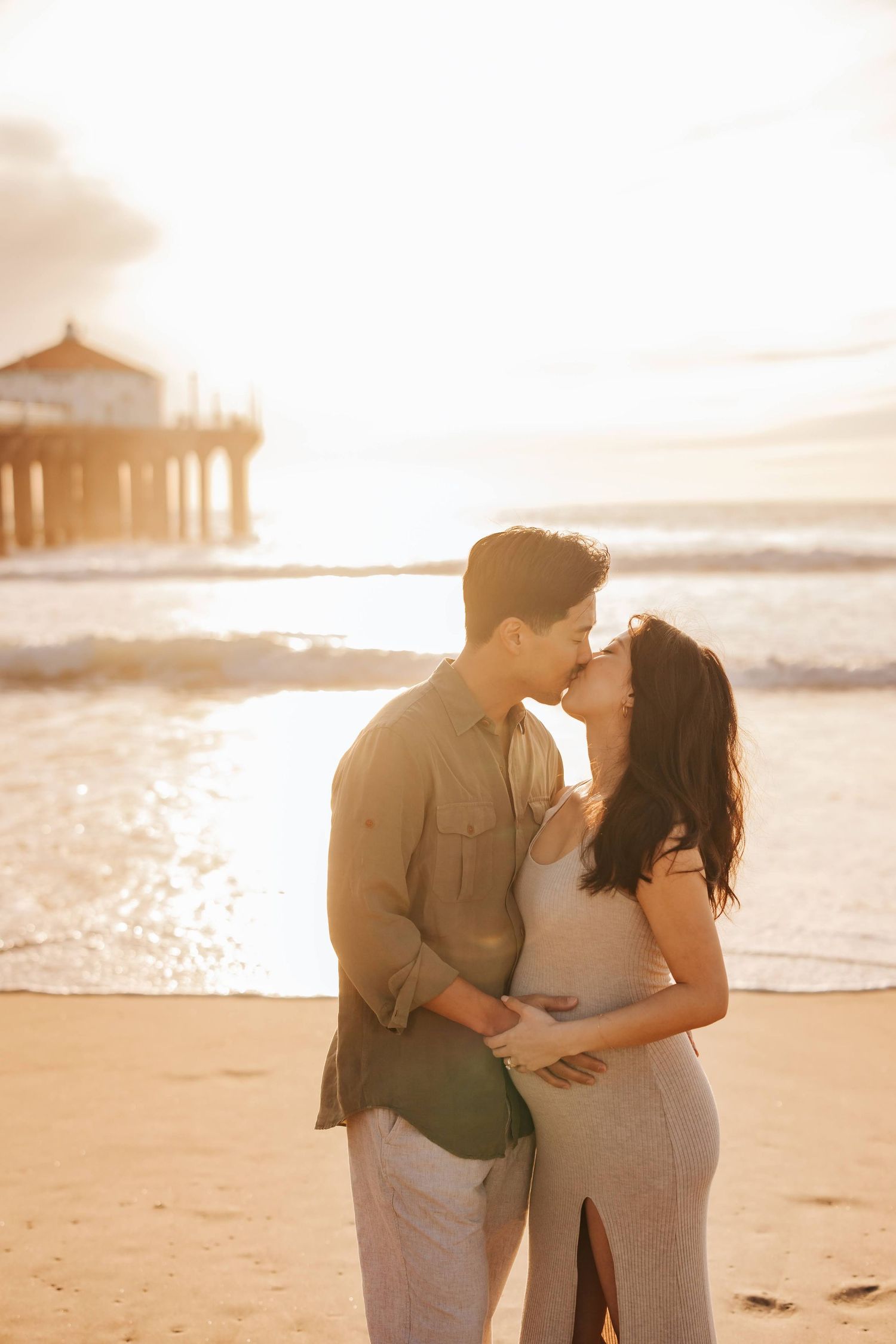 A tender sunset moment between a couple embracing near a beach pier with golden light in the background.
