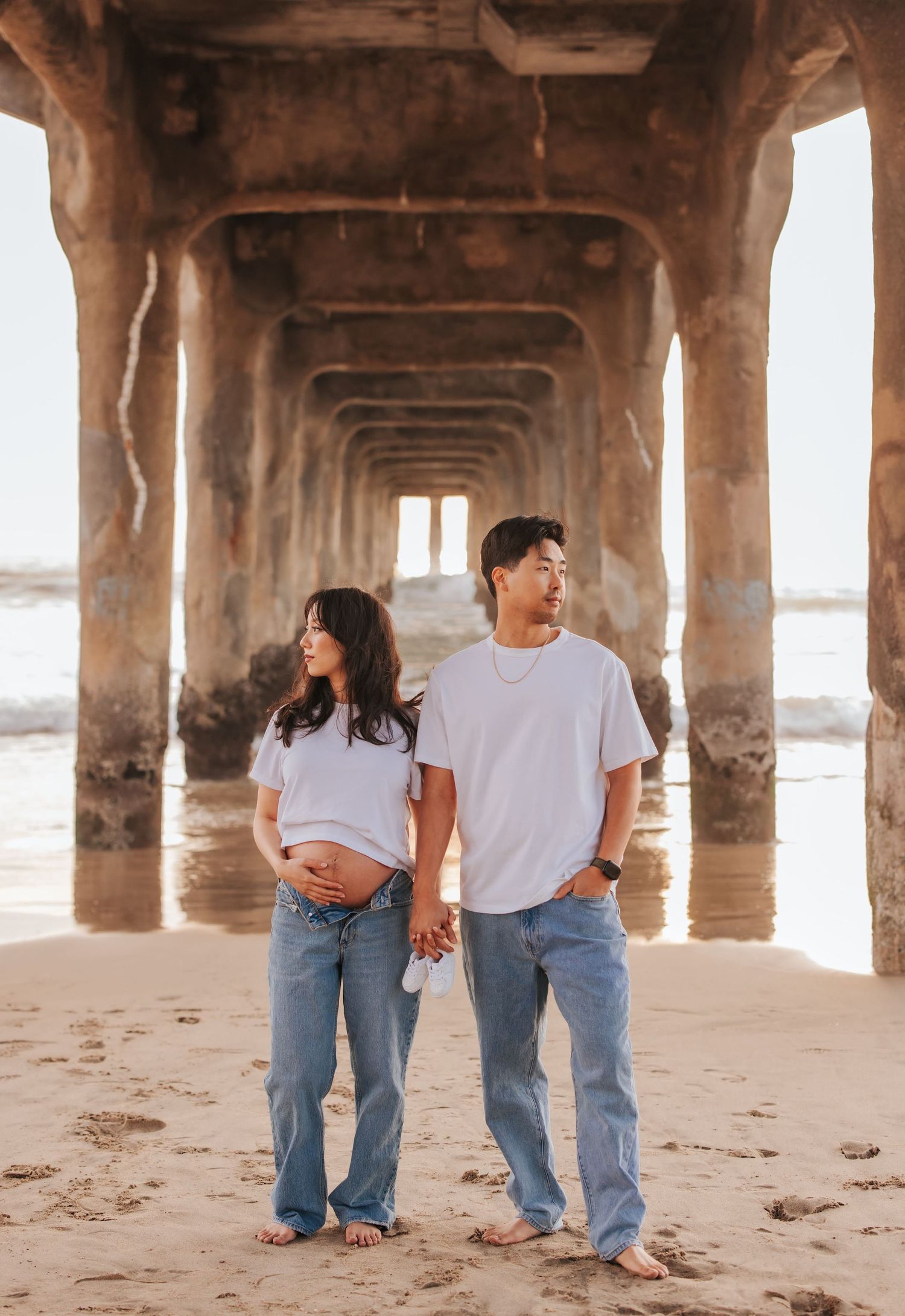 A couple in white shirts and jeans holds hands under a pier at sunset, creating a warm coastal aesthetic.