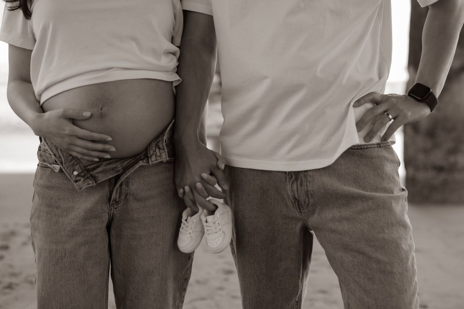 Black and white photo of pregnant belly with a couple's hands holding baby shoes on a beach.