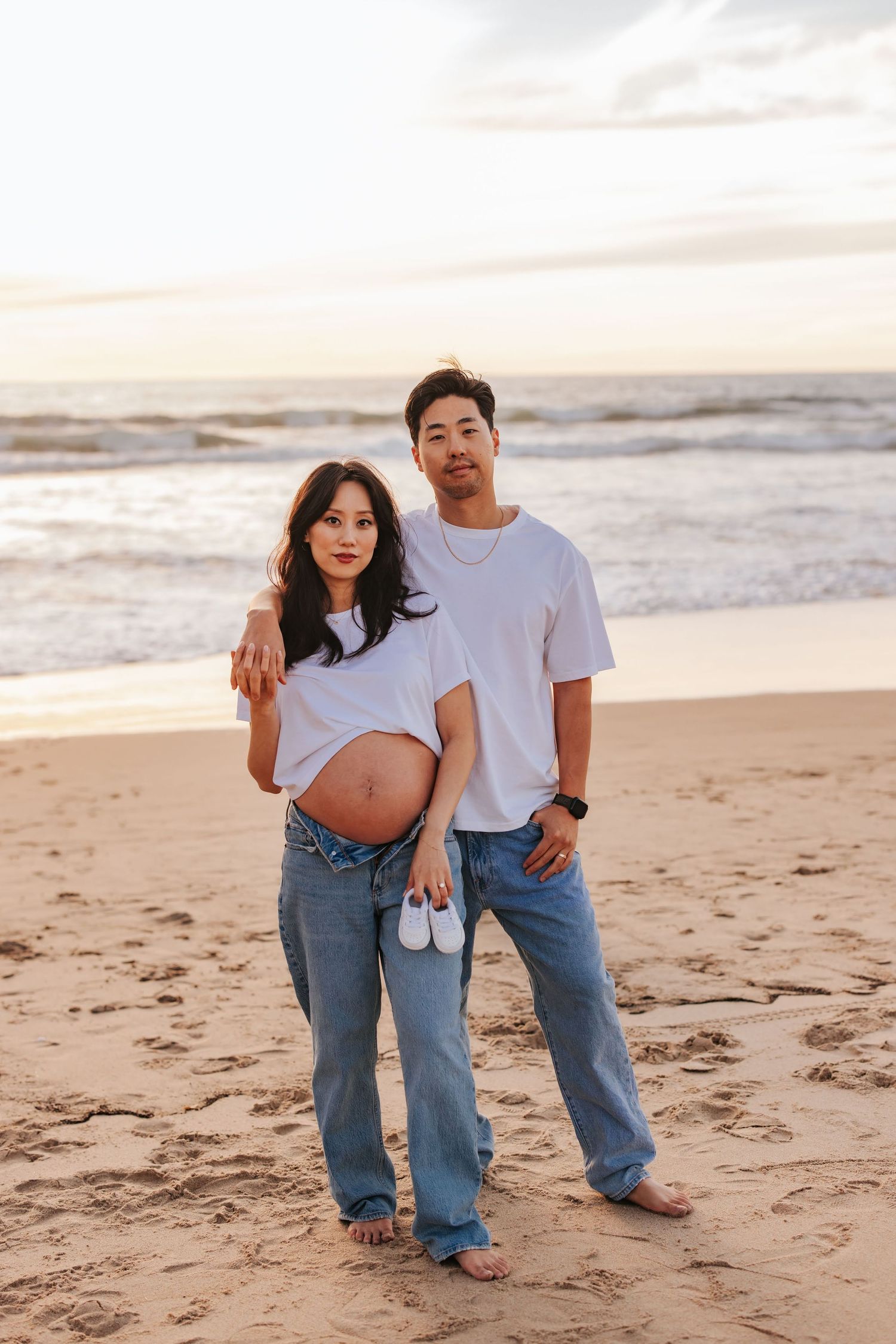 Expectant couple in casual wear holds tiny white baby shoes while standing on a sunset beach.