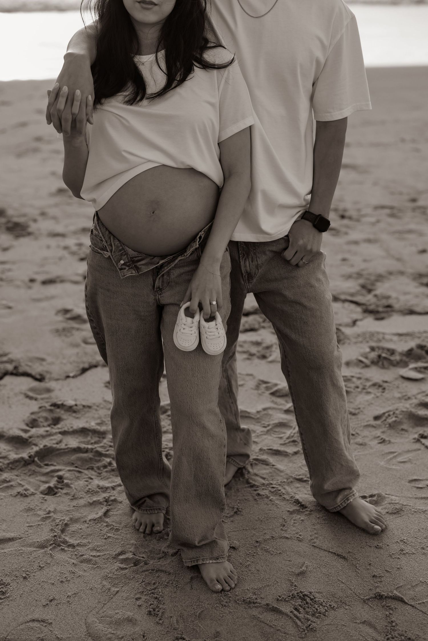 Artistic black and white pregnancy photo showing barefoot couple on beach holding baby shoes.