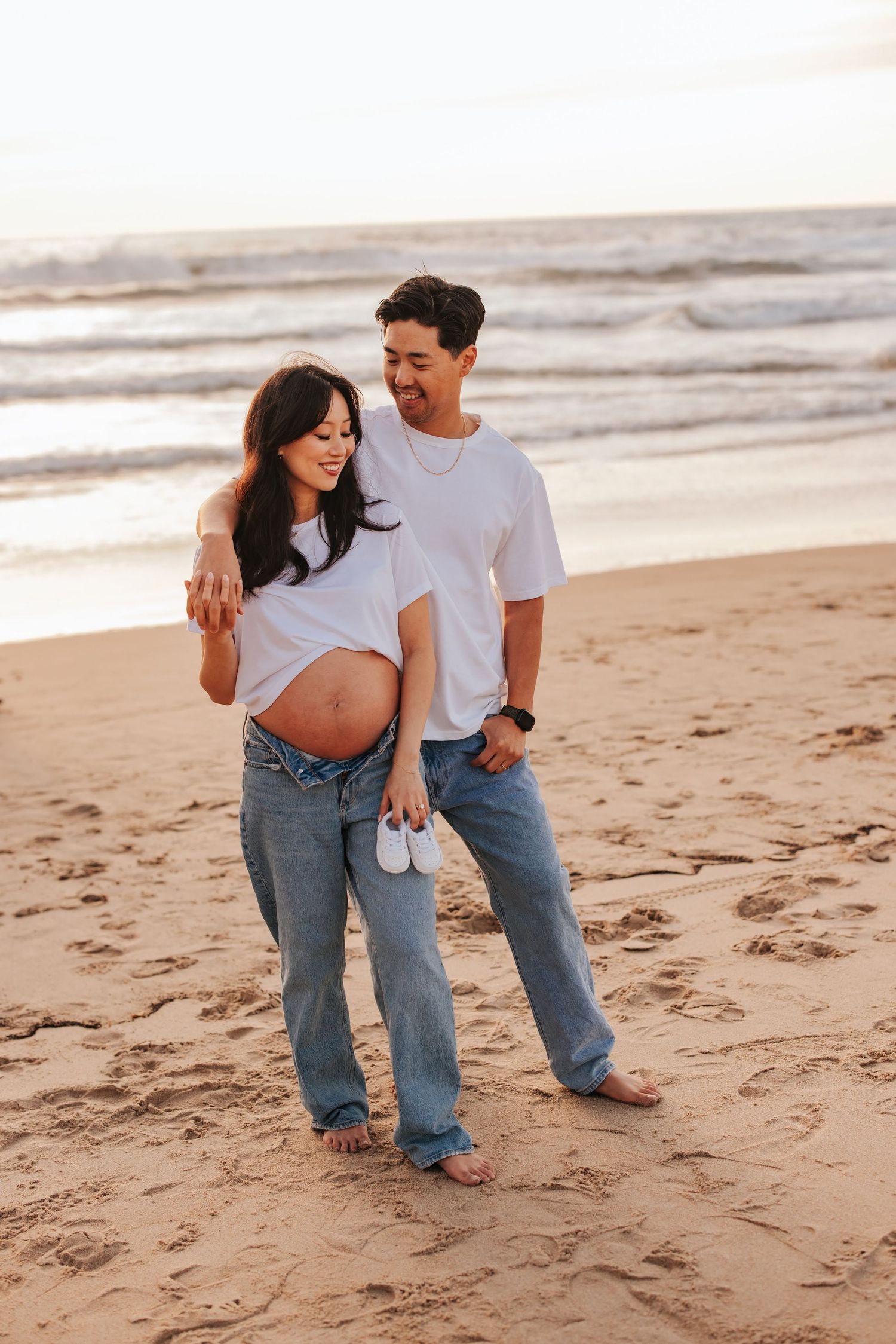 Expectant couple walking together on a sandy beach at sunset while holding small baby shoes.