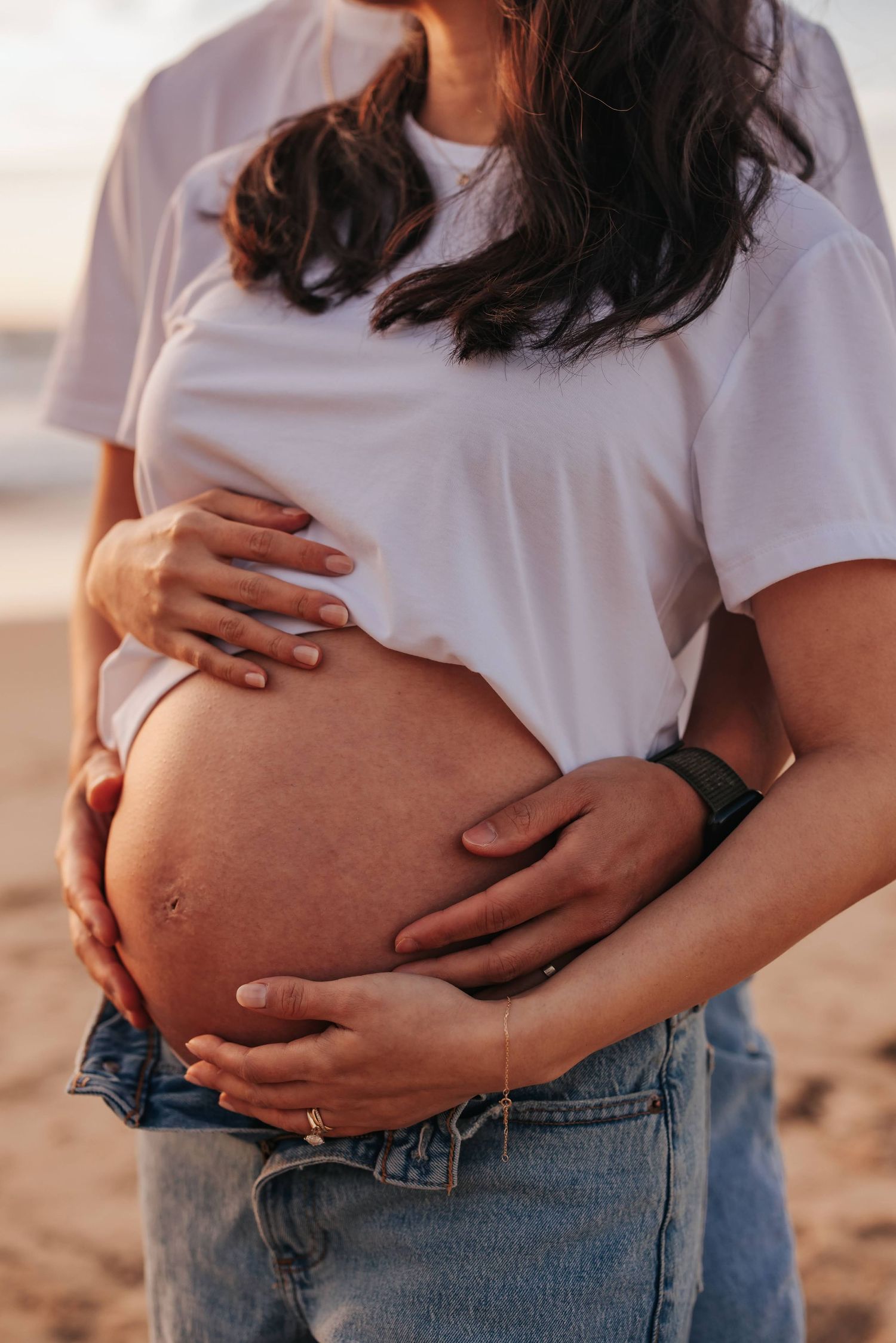 Pregnant woman in white t-shirt and jeans cradles her exposed baby bump on the beach at sunset.