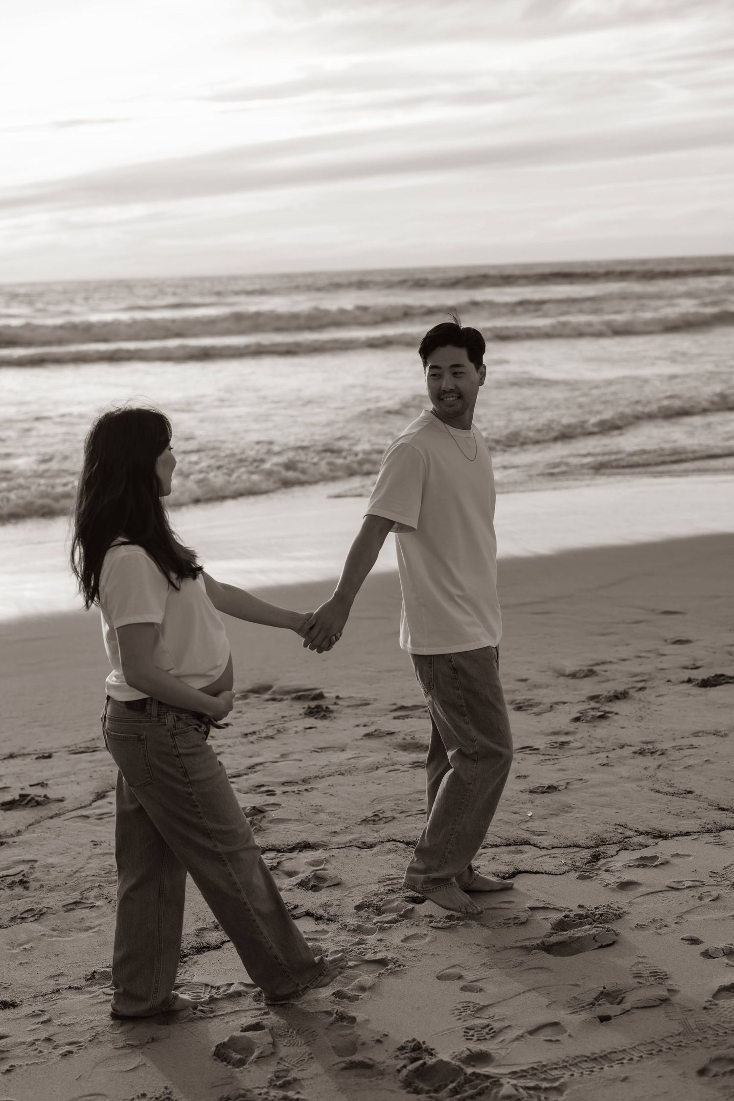 A couple holding hands and walking along a serene beach at sunset in black and white.