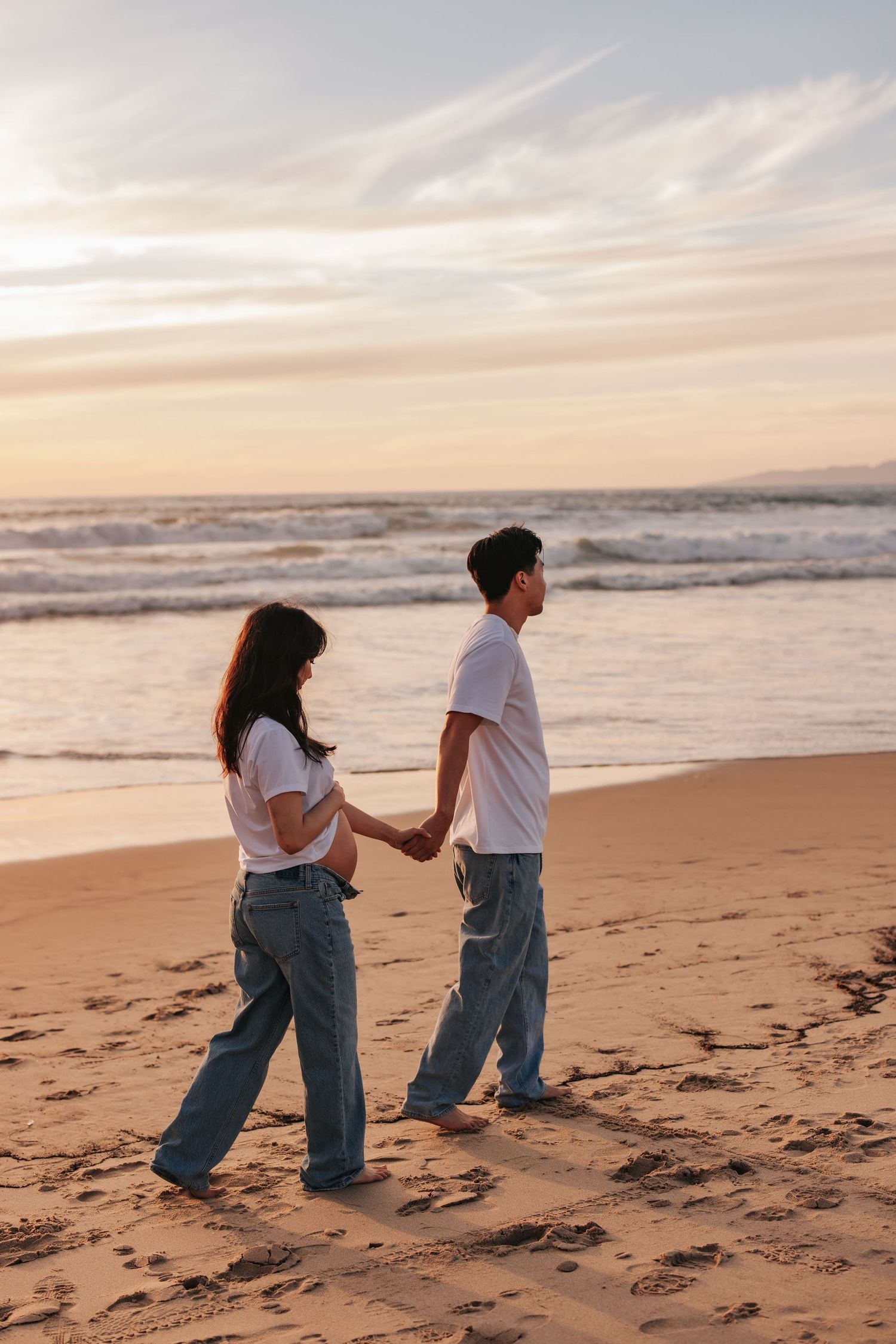 Two people holding hands while walking on sandy beach at sunset with gentle ocean waves in the background.