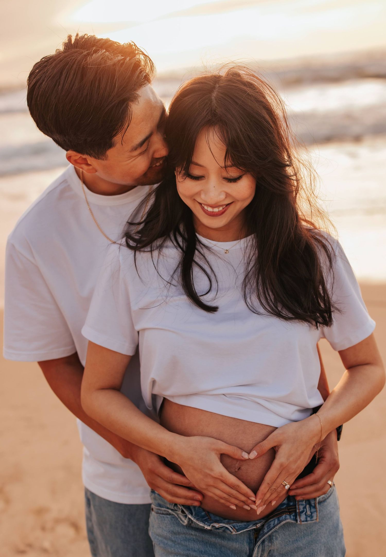 Expecting couple in matching white shirts embrace on the beach, making a heart shape over her pregnant belly.