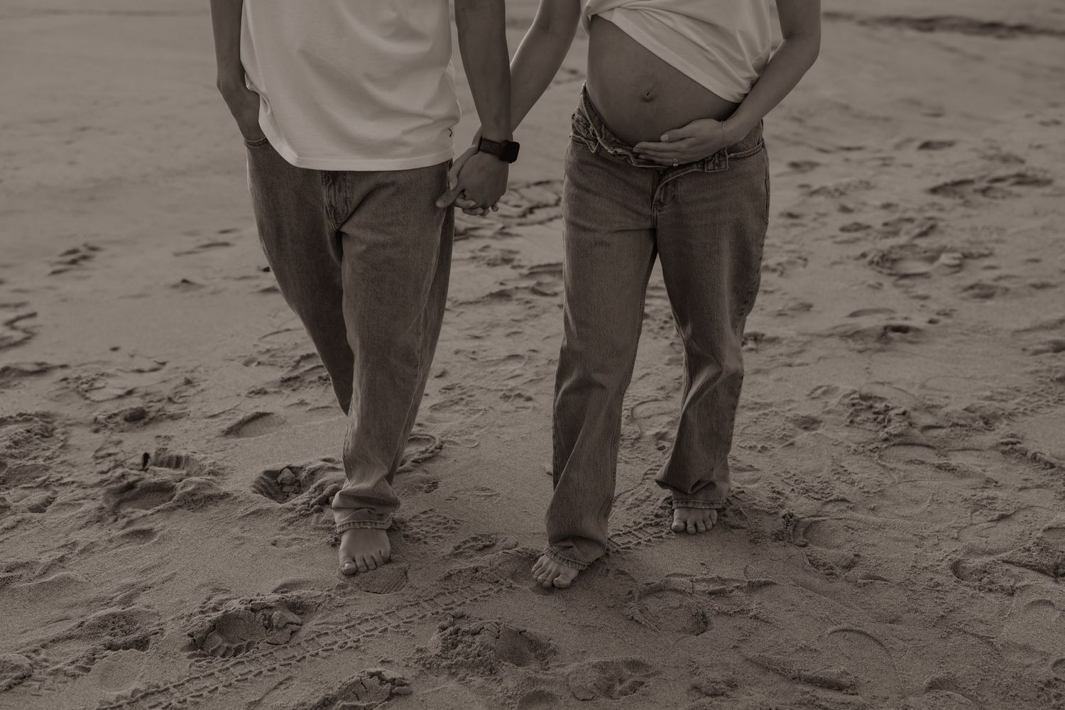 A black and white close-up of two people walking hand in hand on a sandy beach.