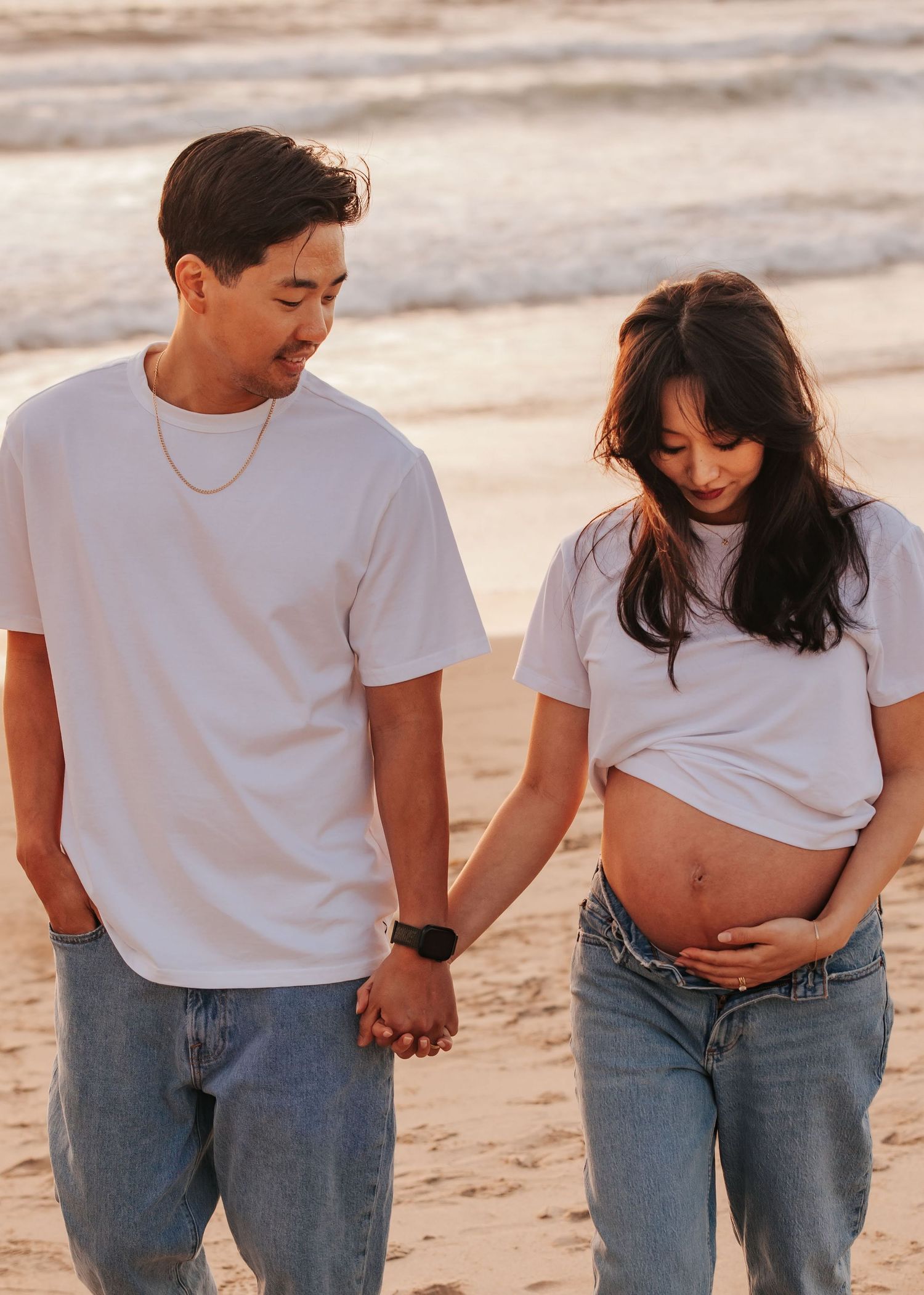 A couple in matching white t-shirts and jeans walk hand in hand on the beach, looking lovingly at an expectant belly.