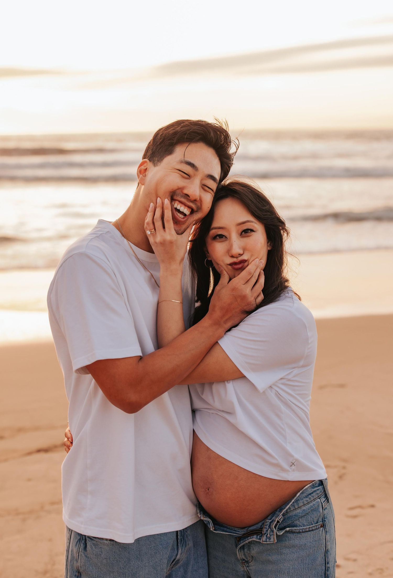 Couple in matching white t-shirts embracing and laughing together on beach at sunset.