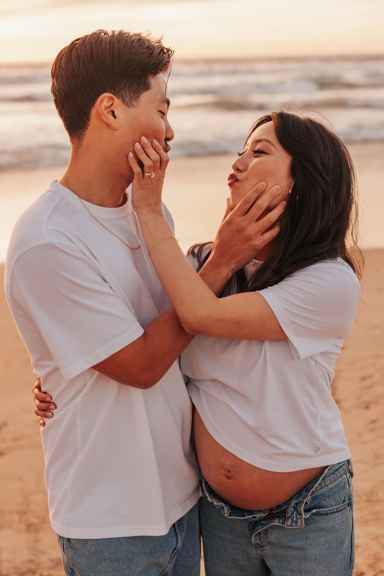 A loving couple in white t-shirts share an intimate moment on the beach at sunset, cupping each other's faces.