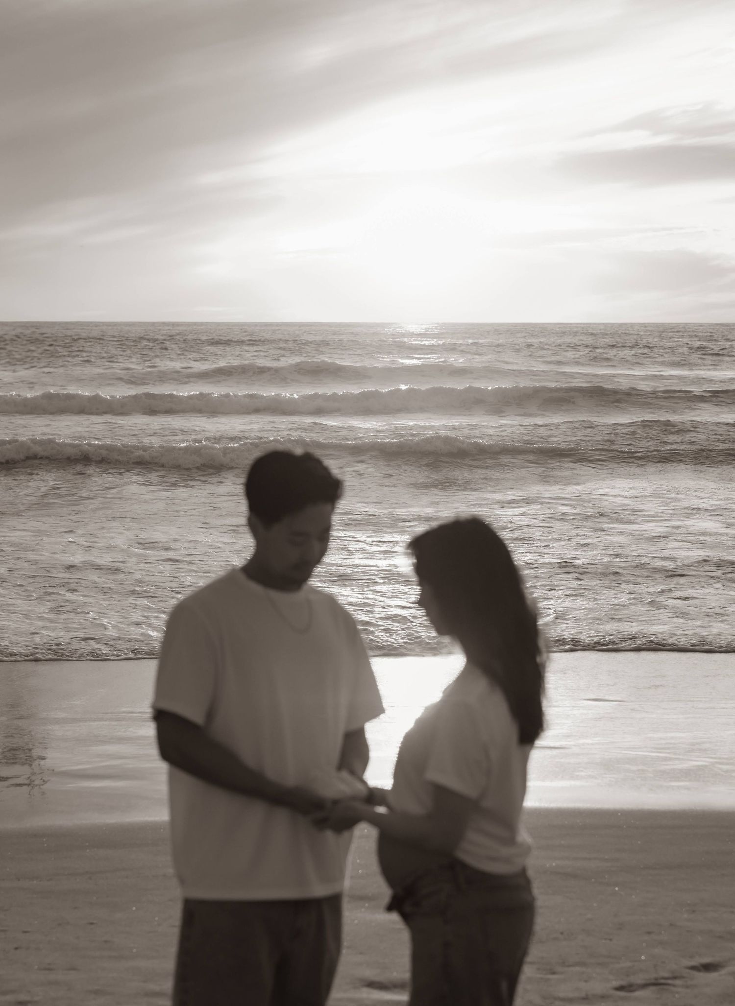 A couple stands in silhouette on a beach at sunset with ocean waves rolling in the background.