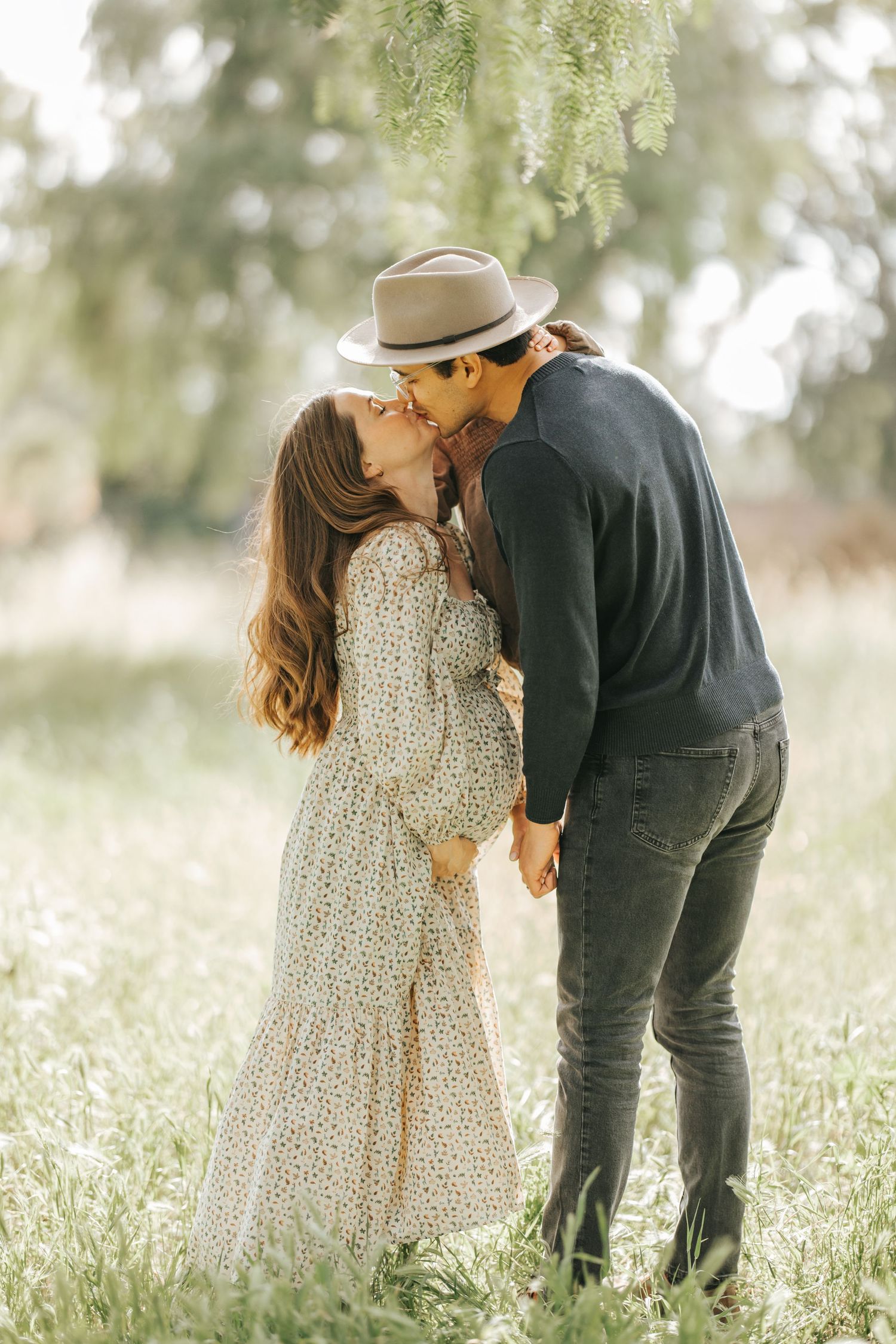 Romantic couple in floral dress and dark sweater share an intimate kiss under willow trees in a dreamy garden setting.