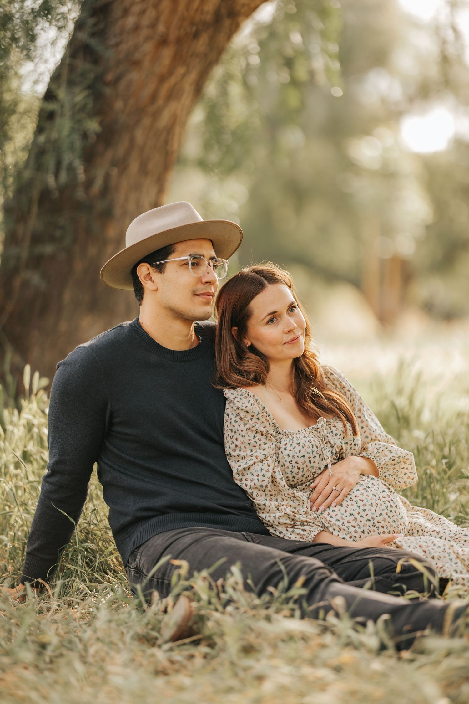 Couple relaxes together under oak tree during golden hour maternity photoshoot.