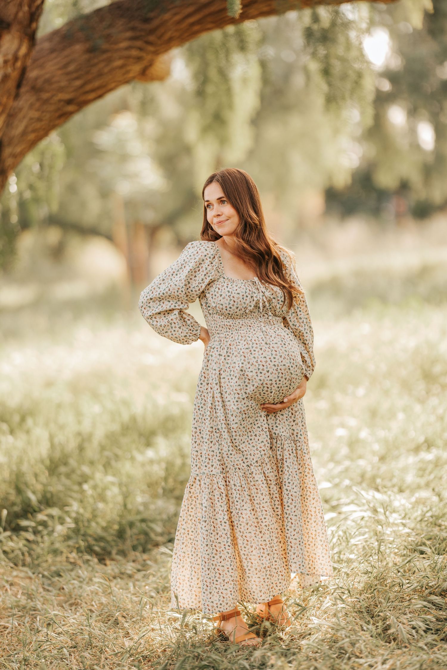 Expectant mother in floral dress poses under tree branches in ethereal lighting.