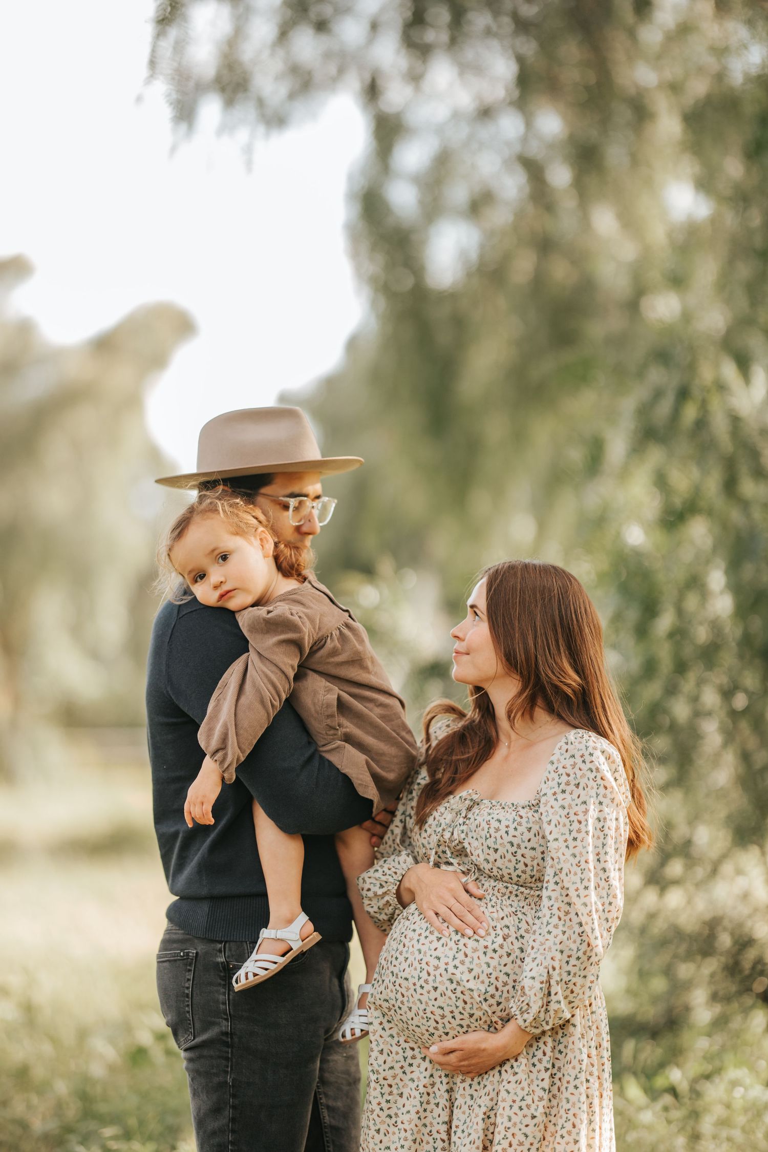 Couple in stylish outfits poses with toddler under willow trees during golden hour.