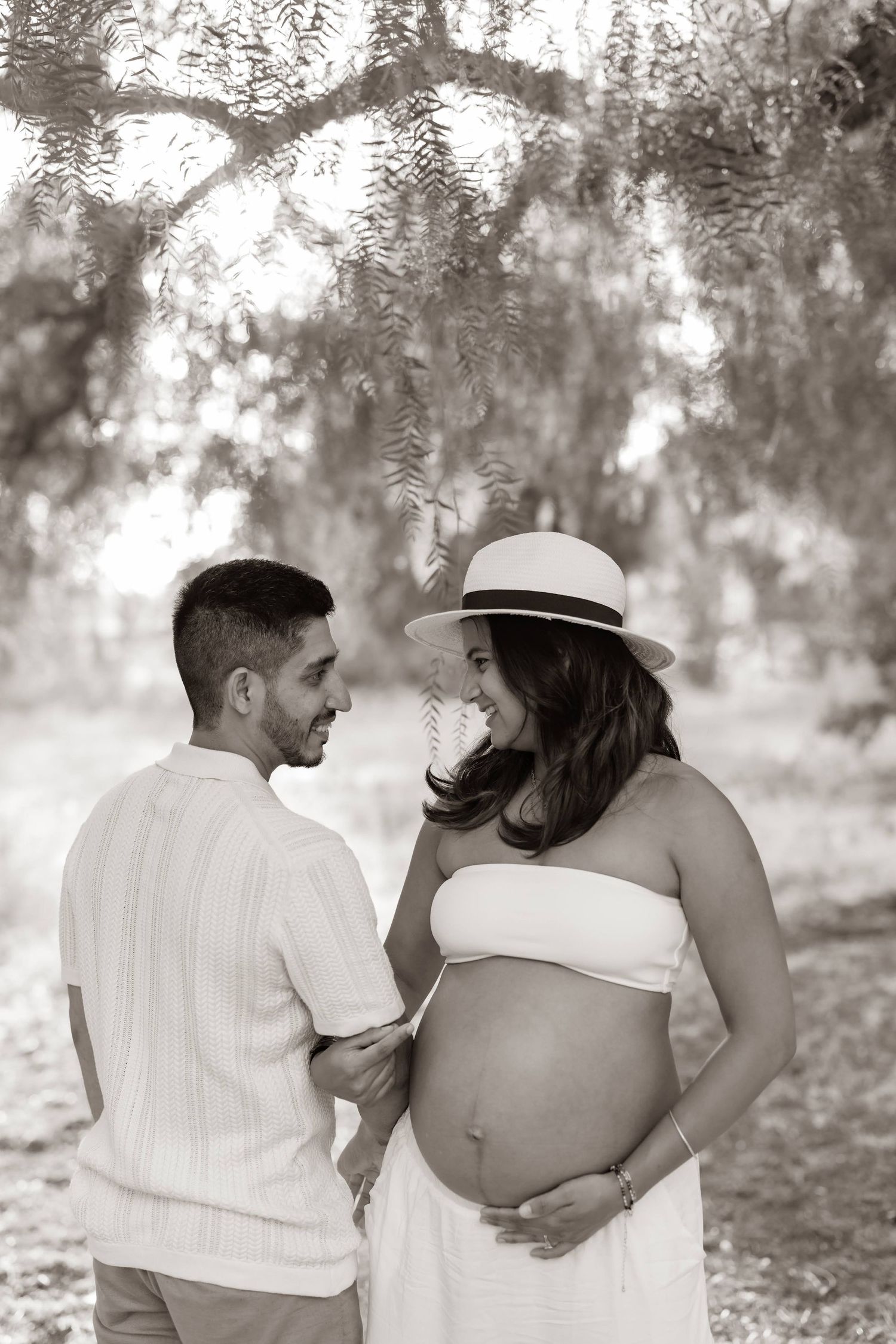 A pregnant individual in a white dress and sun hat stands with their partner outdoors in a dreamy black and white photo.