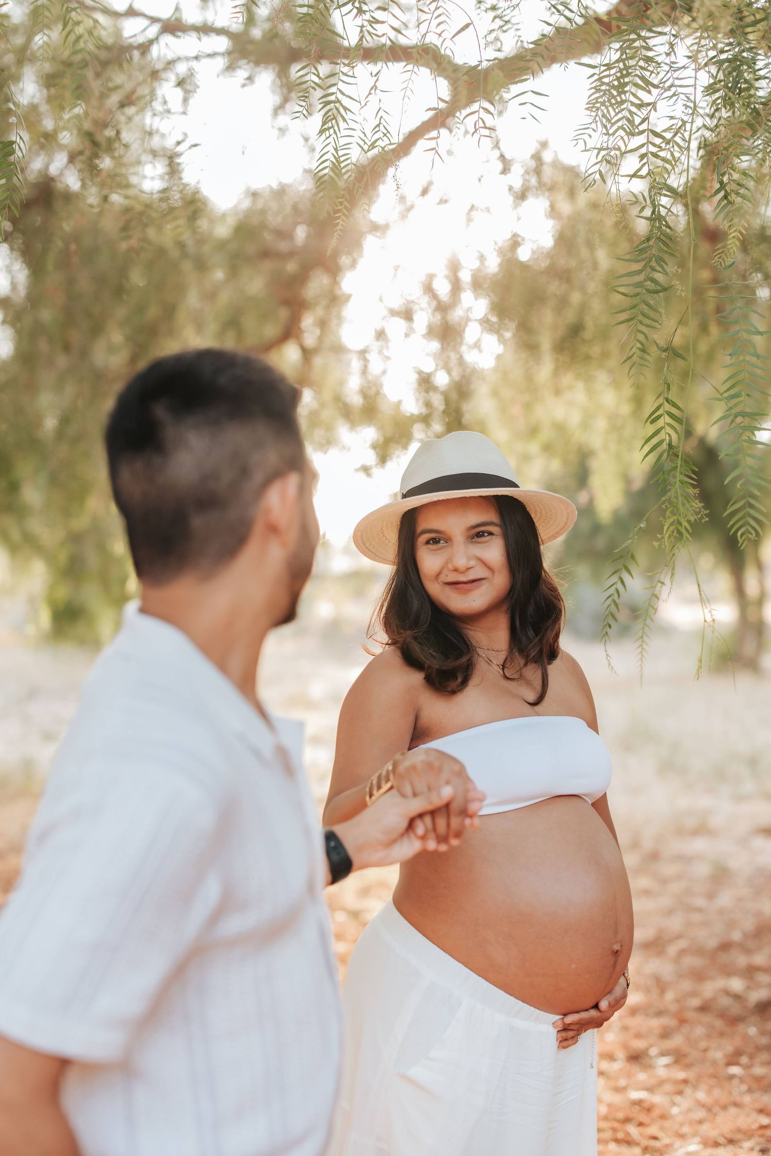 A maternity photo shoot outdoors with a couple in white outfits standing under willow trees at sunset.