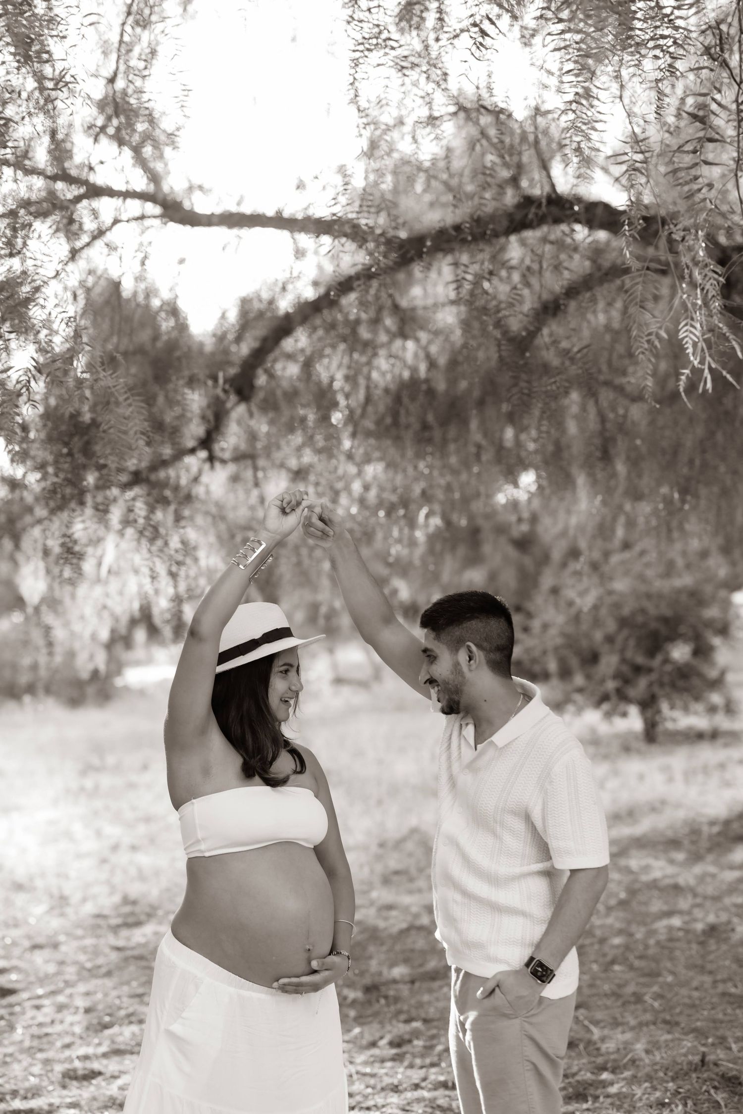 A romantic black and white maternity photo shows an expecting couple sharing a joyful moment under a willow tree.