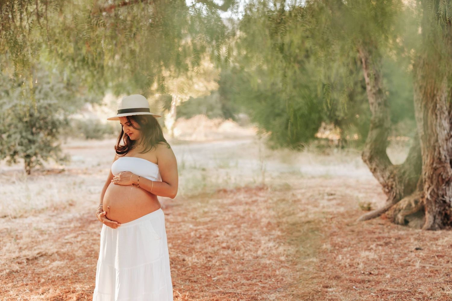 Expectant mother in a flowing white dress and sun hat stands in a dreamy sunlit grove among olive trees.