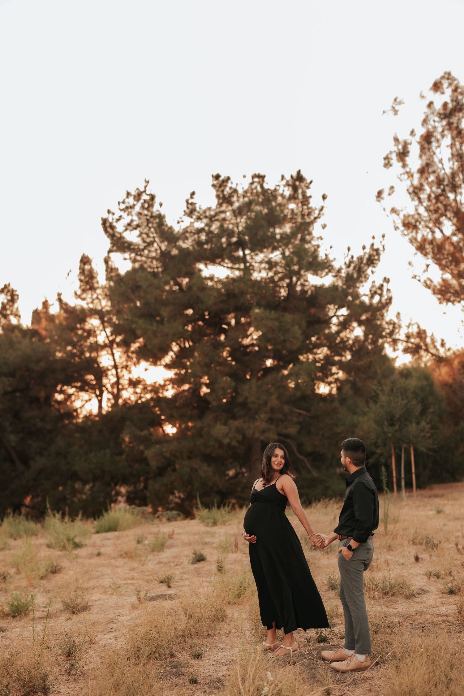 A couple holding hands on a hillside at sunset with silhouetted pine trees in the background.