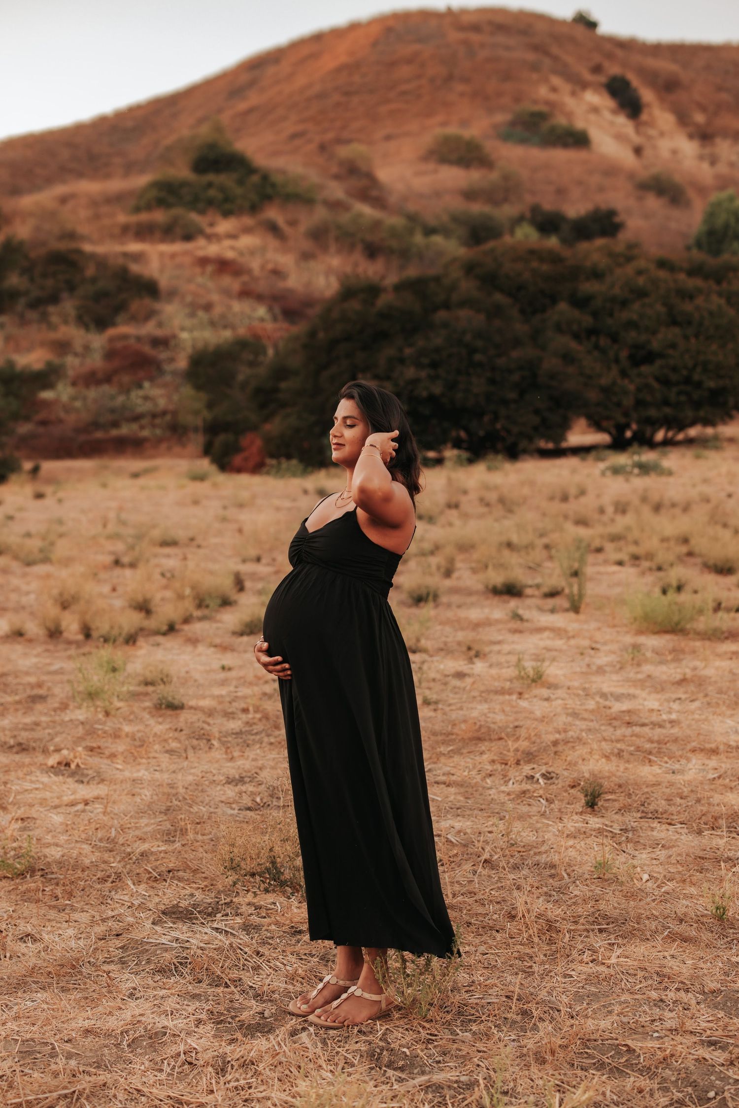 A pregnant woman in a flowing black maxi dress poses barefoot against a backdrop of rustic golden hills and trees.