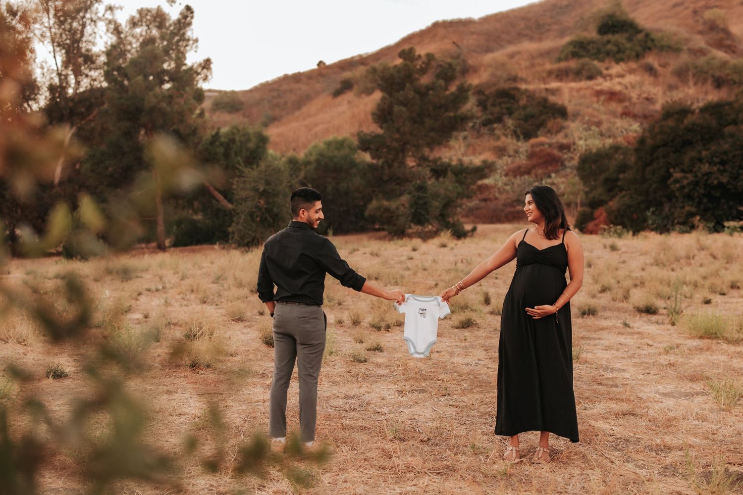 Two people hold a small white garment while standing in a golden brown field with hills in the background at sunset.