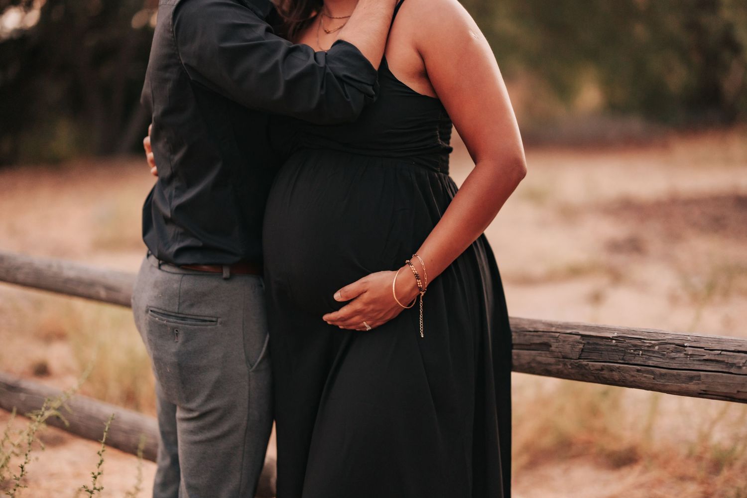 Two people embracing in black clothing stand by a rustic wooden fence during a warm sunset photoshoot.