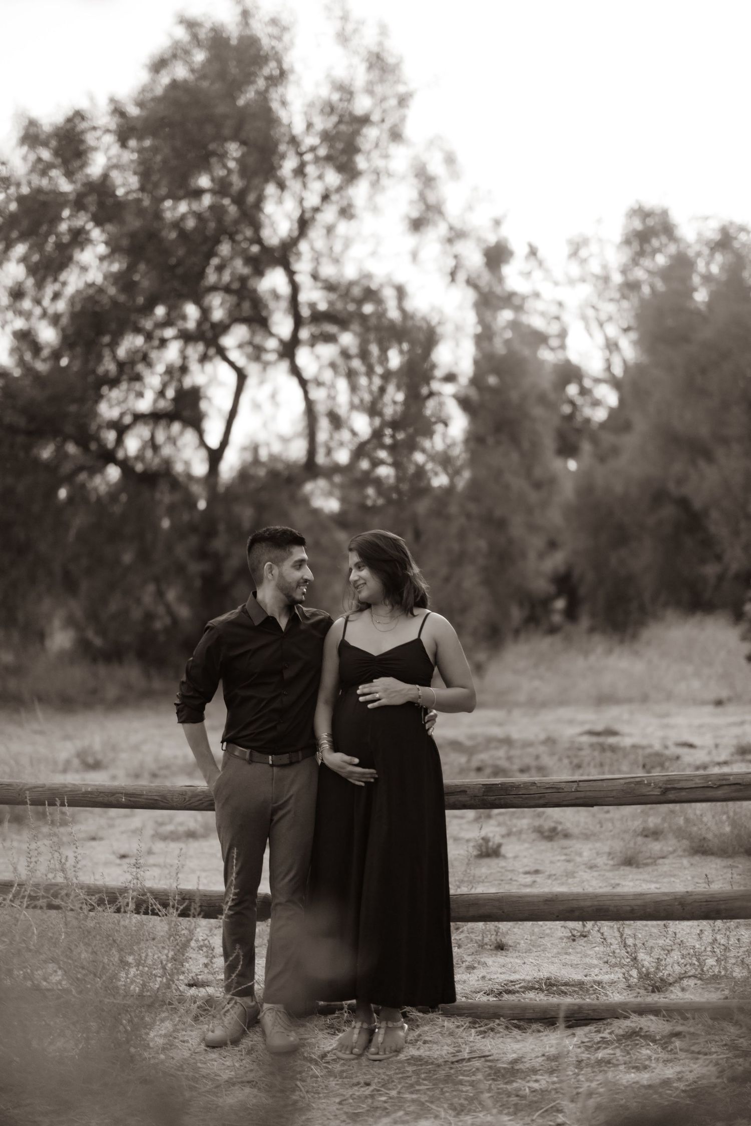 A couple shares an intimate moment in black and white by a rustic wooden fence as the sun sets behind trees.