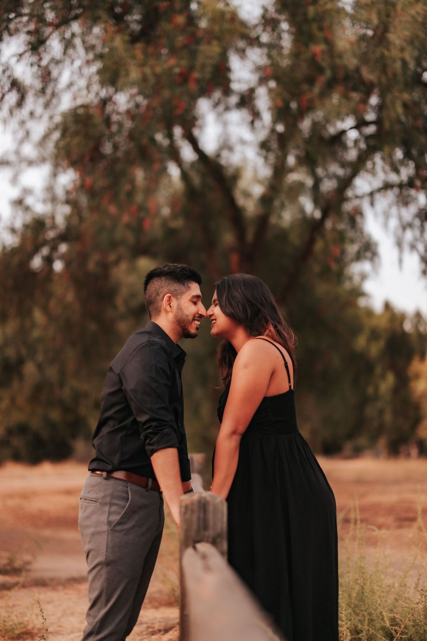 Romantic couple in formal attire share an intimate moment beside a wooden fence at sunset in an autumn setting.