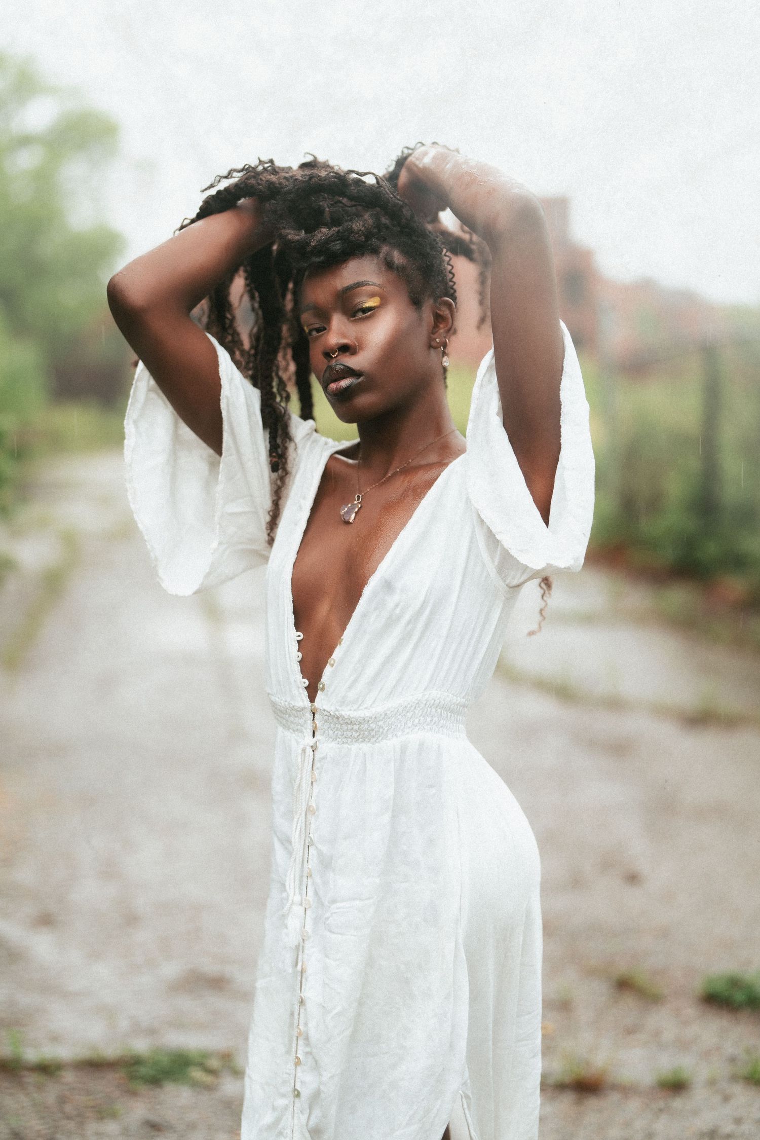 Black woman in white deep-V boho dress with locs raised overhead posing on misty outdoor path in Cincinnati