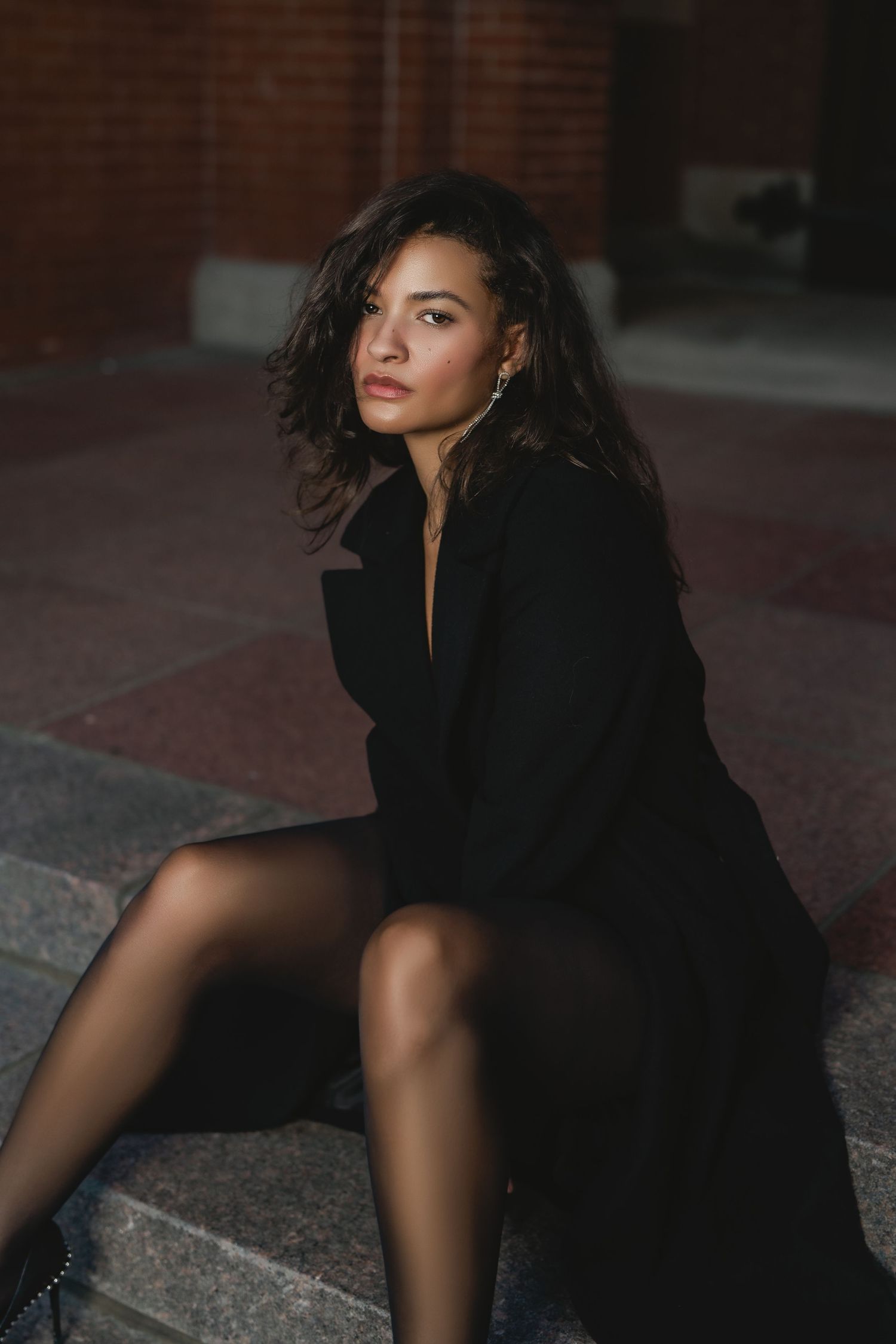 Young woman in black coat sitting on steps outdoors with brick wall background in moody lighting in Cincinnati