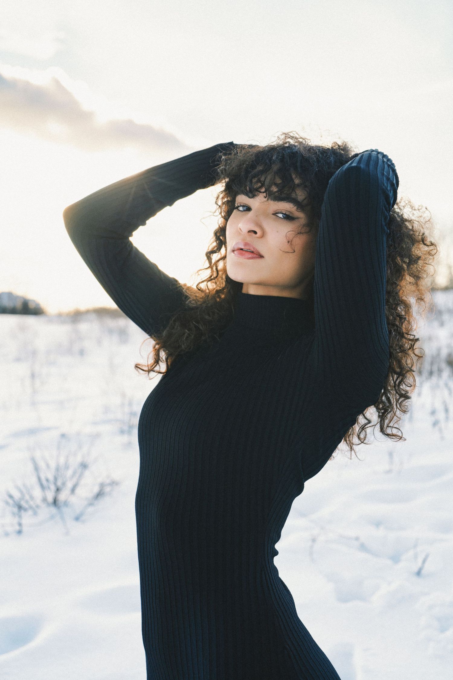 Young woman with curly hair in black turtleneck sweater dress posing in snowy winter landscape with sunlight behind her in Cincinnati