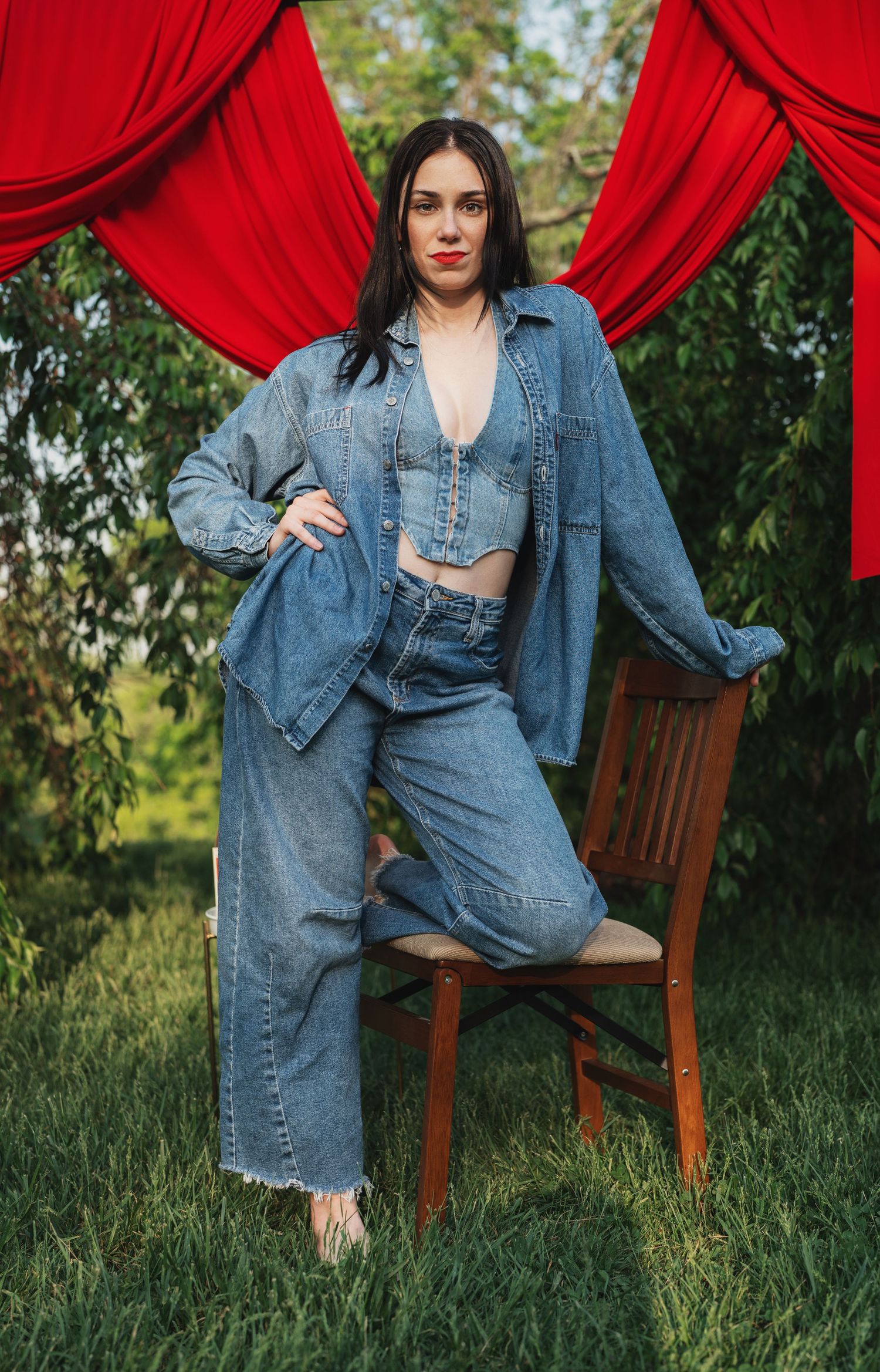 Woman in a jean jacket, pants and jean blouse with a knee on a chair in a wooded area with red drapes
