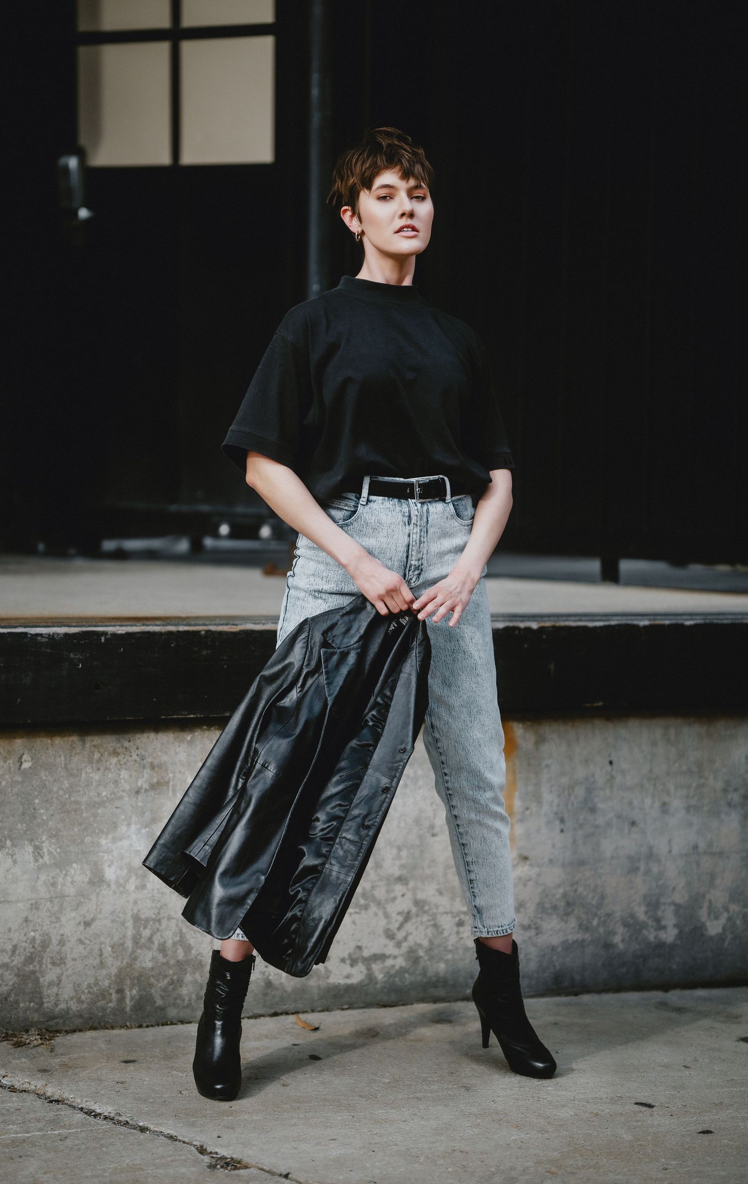 Woman in black tee, light wash jeans, and ankle boots holding leather jacket in urban setting in Cincinnati