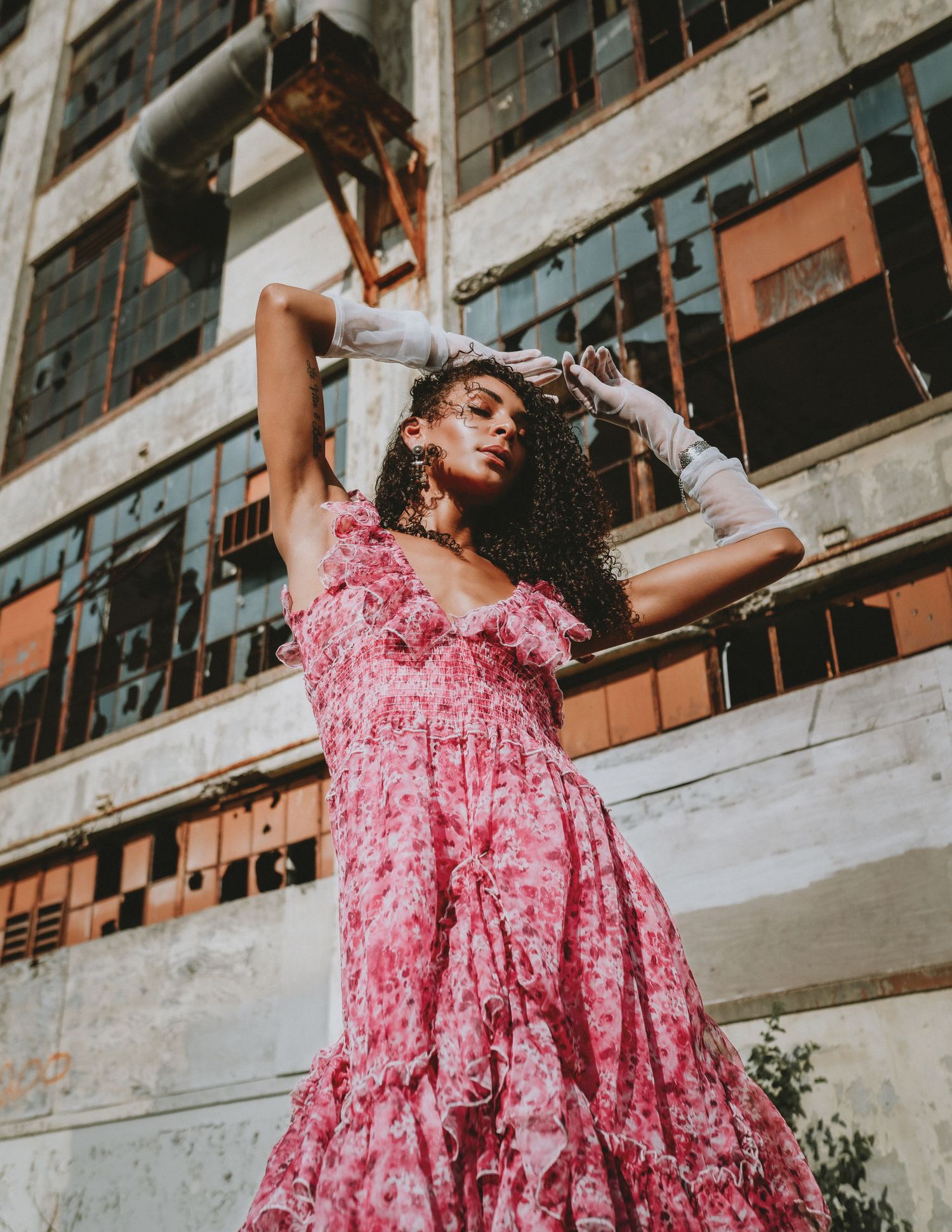 Woman in pink floral midi dress with white gloves posing dramatically in front of industrial building for her fashion shoot in Cincinnati