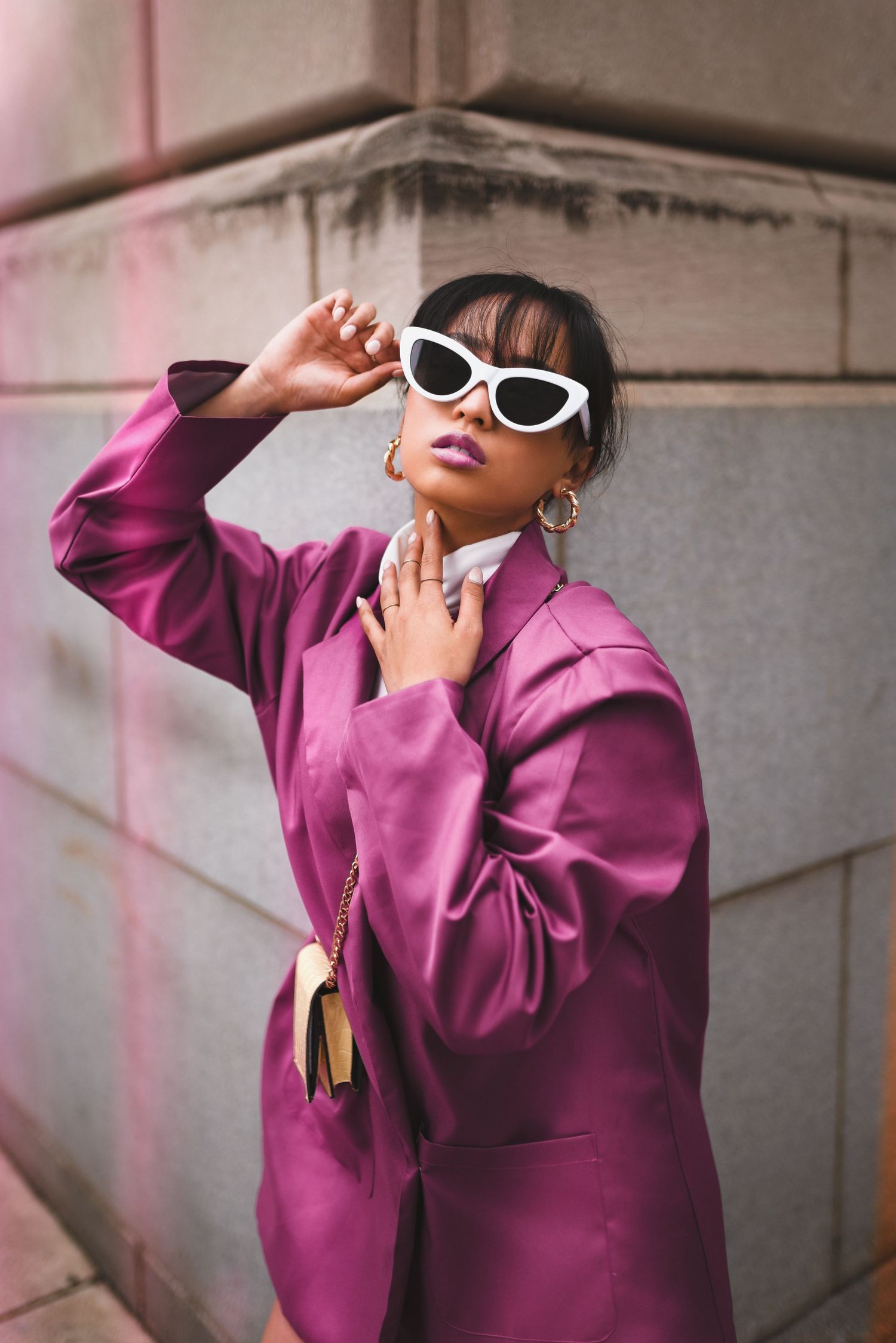 Stylish woman in magenta oversized blazer wearing white cat-eye sunglasses posing against stone wall in Cincinnati
