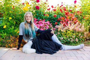 Blonde girl with teal highlights sits smiling in front of vibrant colorful flower garden on brick patio.