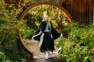 Blonde woman in black cape and skirt posing in a circular garden gate surrounded by lush green foliage.