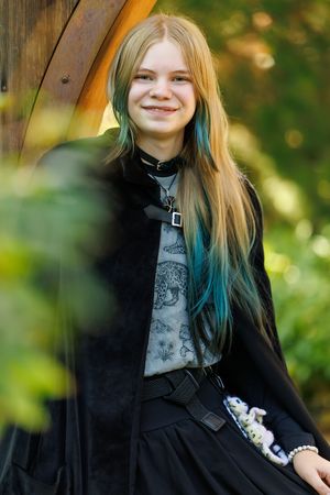 Smiling teenage girl with long blonde and teal hair wearing black gothic outfit outdoors.