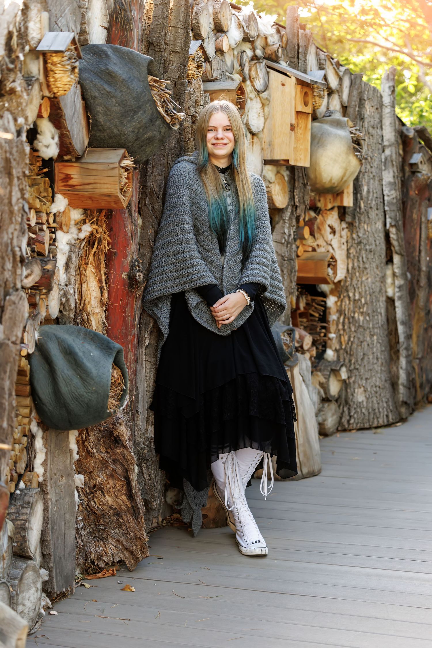 Young woman in gray knit poncho and black skirt smiling against rustic log wall with birdhouses.
