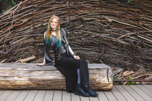 Woman with teal-highlighted hair sits on a log bench against a rustic twig wall backdrop, wearing a black outfit.