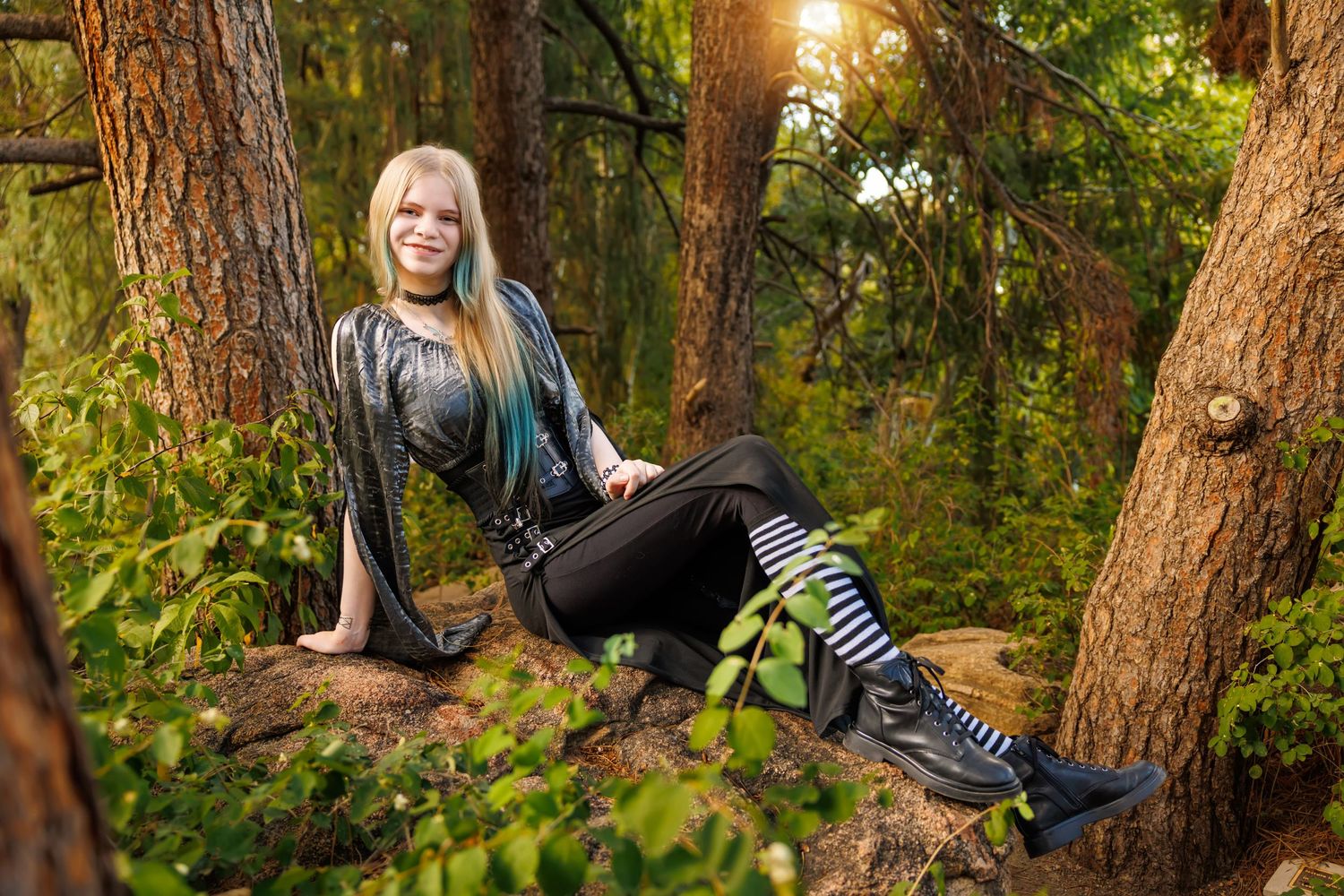 Smiling blonde woman in black outfit with striped boots sitting on rocks in a sunlit forest setting.