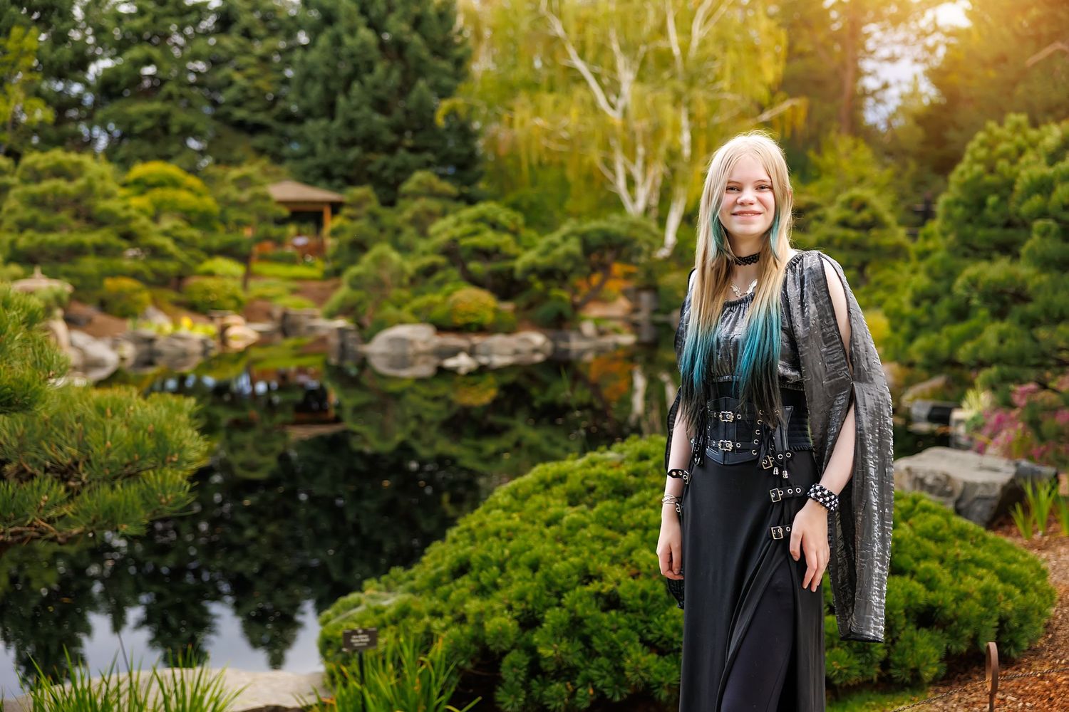 Young woman with teal-tipped blonde hair poses in a Japanese garden with pond and pagoda.