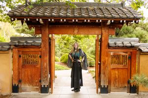 Person in black kimono standing under a traditional Japanese wooden gate surrounded by lush greenery.